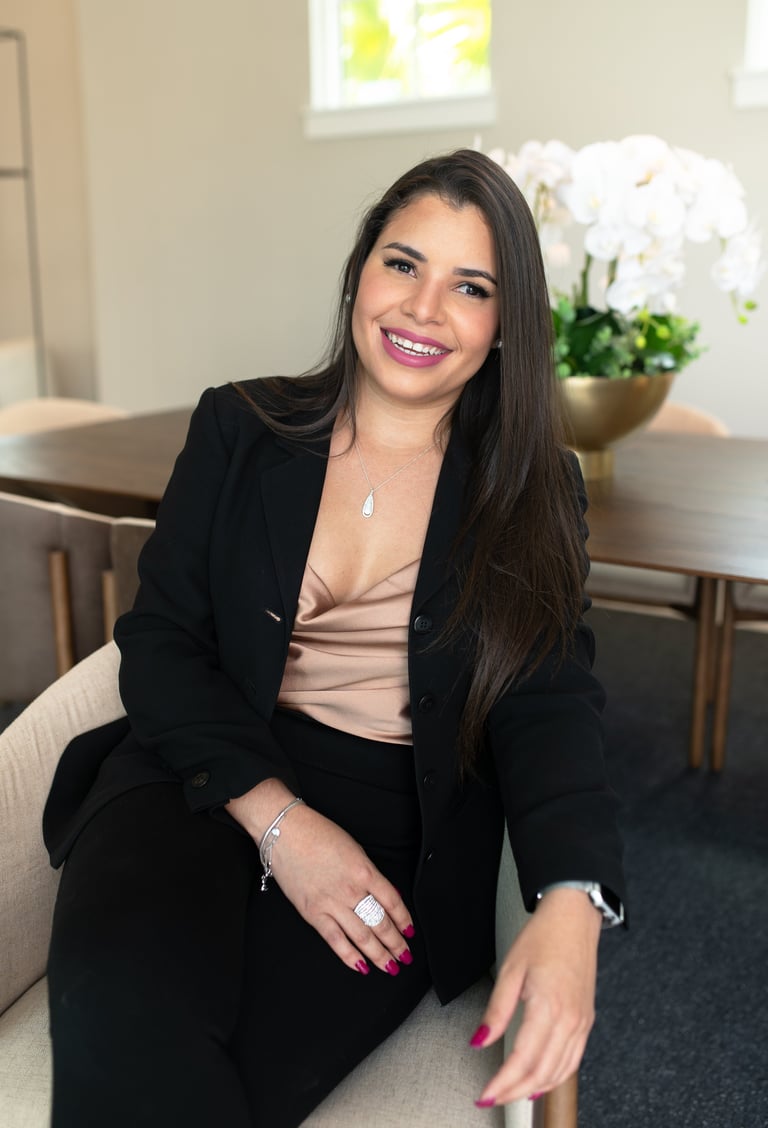 Professional woman with long dark hair wearing a black blazer and beige top, smiling at camera while seated in an office setting with flowers in background